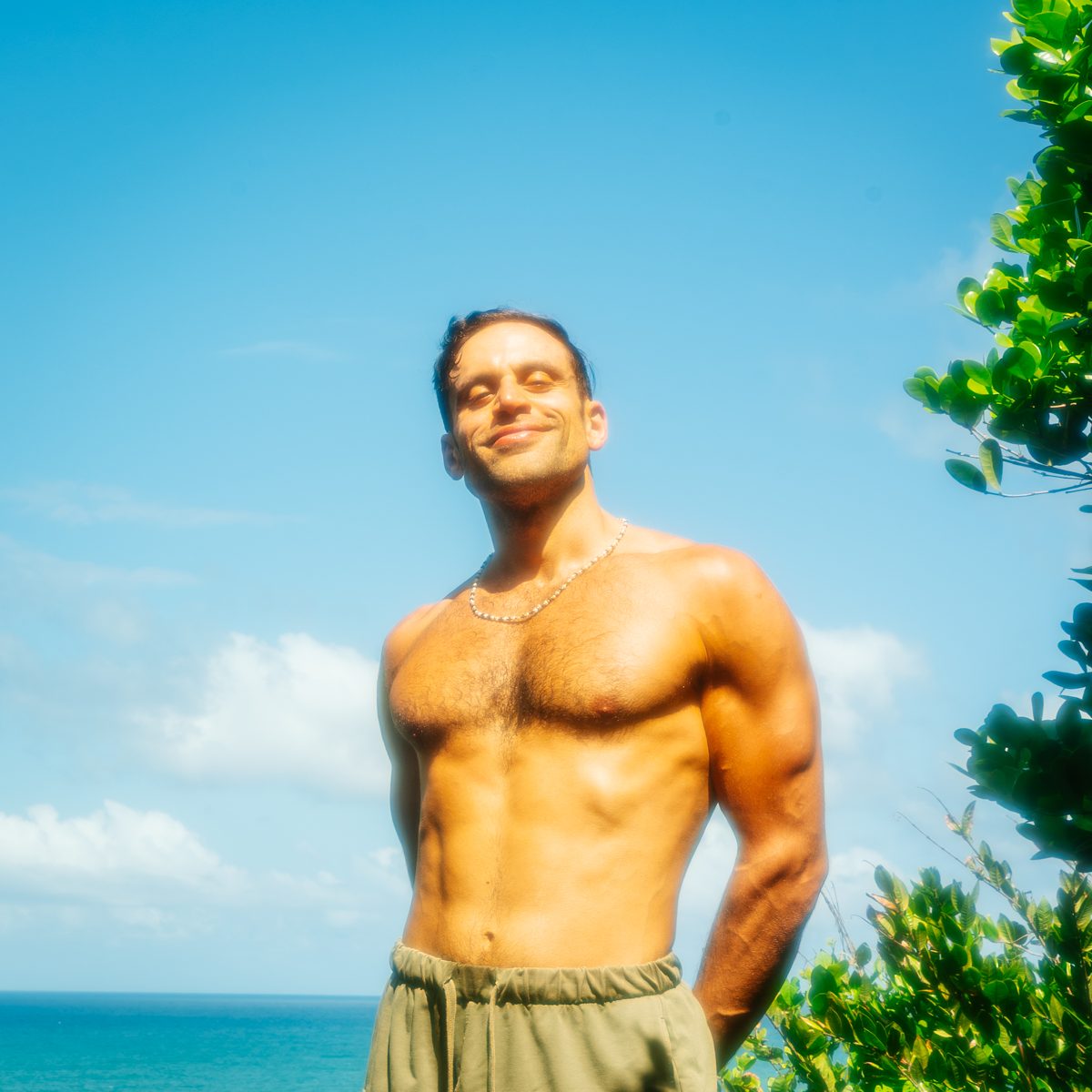 Sid shirtless on a cliff above the sea, eyes closed to the sun